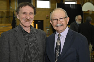 Two senior men smiling at a community event, dressed in formal attire with a gymnasium setting in the background.