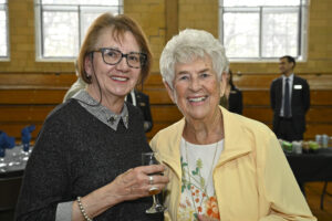 Two elderly women smiling at a social event, one holding a glass of wine. The background features a table with drinks and two men in suits. The event appears to take place in a well-lit room with large windows and wooden walls.