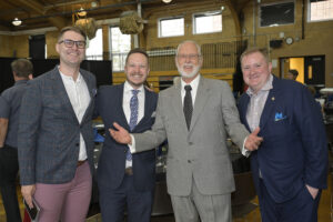 Four men in suits smiling and posing for a group photo at an indoor event with a yellow brick wall and chairs in the background.