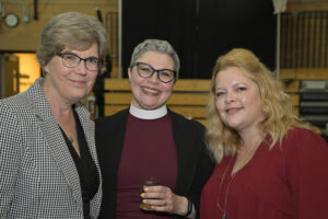 Three women smiling at a social event, one of them dressed in clergy attire. The image captures a moment of camaraderie and camaraderie. The background suggests the setting is inside a community hall or recreational facility. Keywords: social event, camaraderie, women, clergy attire, community hall.