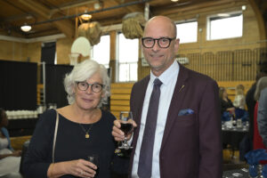 Two elderly people enjoying a formal event in a gymnasium, holding glasses of wine, dressed in professional attire