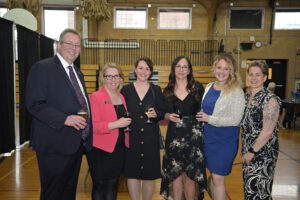 A group of six well-dressed people posing and enjoying drinks at an indoor social event. The setting appears to be a gymnasium with wooden bleachers and high ceilings. Four women, one in a pink blazer, another in a black dress, a third in a floral dress, and the fourth in a blue dress with a silver jacket, stand in the middle, while a man in a suit and tie, and a woman in a patterned dress with a sleeve tattoo, stand on either side.