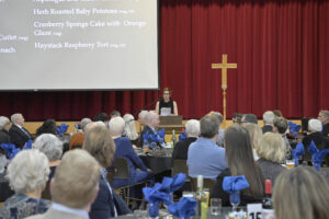 Speaker addressing the audience at a formal event with a cross in the background and menu projected on screen.