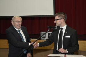 Two men in suits shaking hands at an award ceremony, one standing behind a microphone and a podium, with a red curtain in the background.