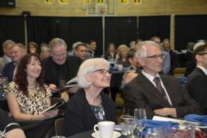 Audience enjoying a formal event, with smiles and attention on speakers, seated at tables with drinks and decorations in a well-lit venue.