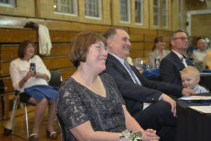 A senior couple smiling and enjoying a public event in a gymnasium with other attendees in the background, including a young child and a woman with a camera.