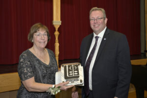 Two people standing together at an award ceremony, one holding a 2023 Distinguished Faculty Award plaque. They are dressed formally with a red curtain in the background.