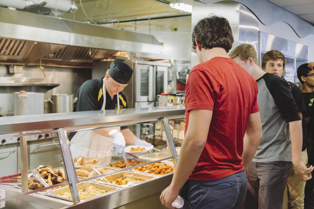 Students line up at a cafeteria serving station where a chef, wearing a black hat and apron, prepares plates of hot food, including chicken, macaroni, and hash browns. The kitchen features stainless steel appliances and overhead ventilation systems.