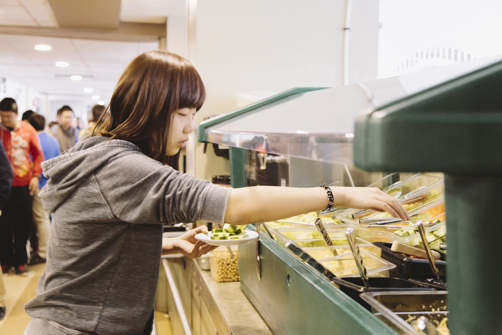 Young woman selecting food at a salad bar in a bustling cafeteria.