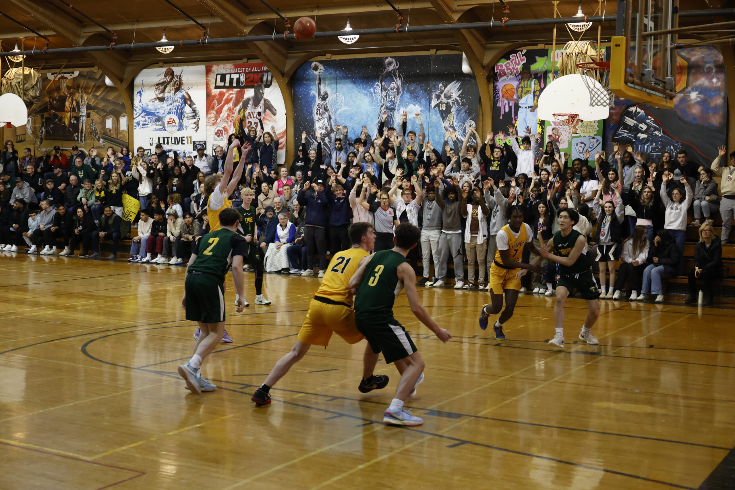 High School Basketball Game: Exciting action during a high school basketball game with players in green and yellow uniforms. The packed gymnasium is filled with enthusiastic fans cheering from the bleachers. Colorful artwork and banners adorn the walls, enhancing the energetic atmosphere.